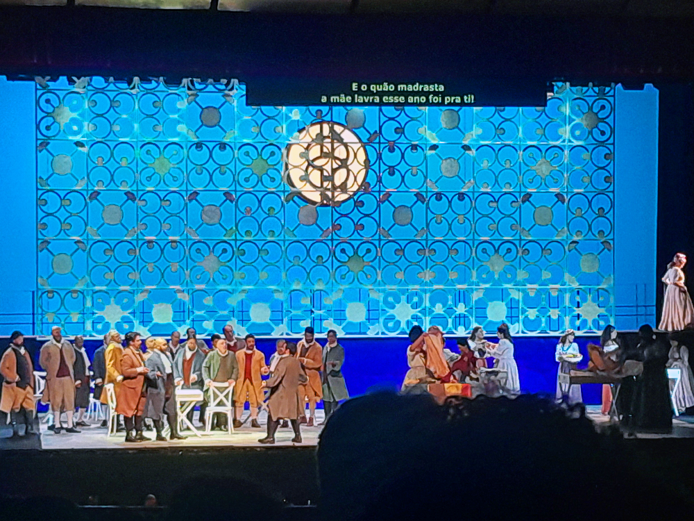 Grande grupo de artistas ocupa o palco, todos em figurinos históricos. O fundo azul traz padrões circulares e elemento luminoso central que lembra vitral. Alguns personagens conversam, outros se movimentam entre mesas, criando sensação de comunidade e conflito. Acima, legenda em português acompanha o libreto.