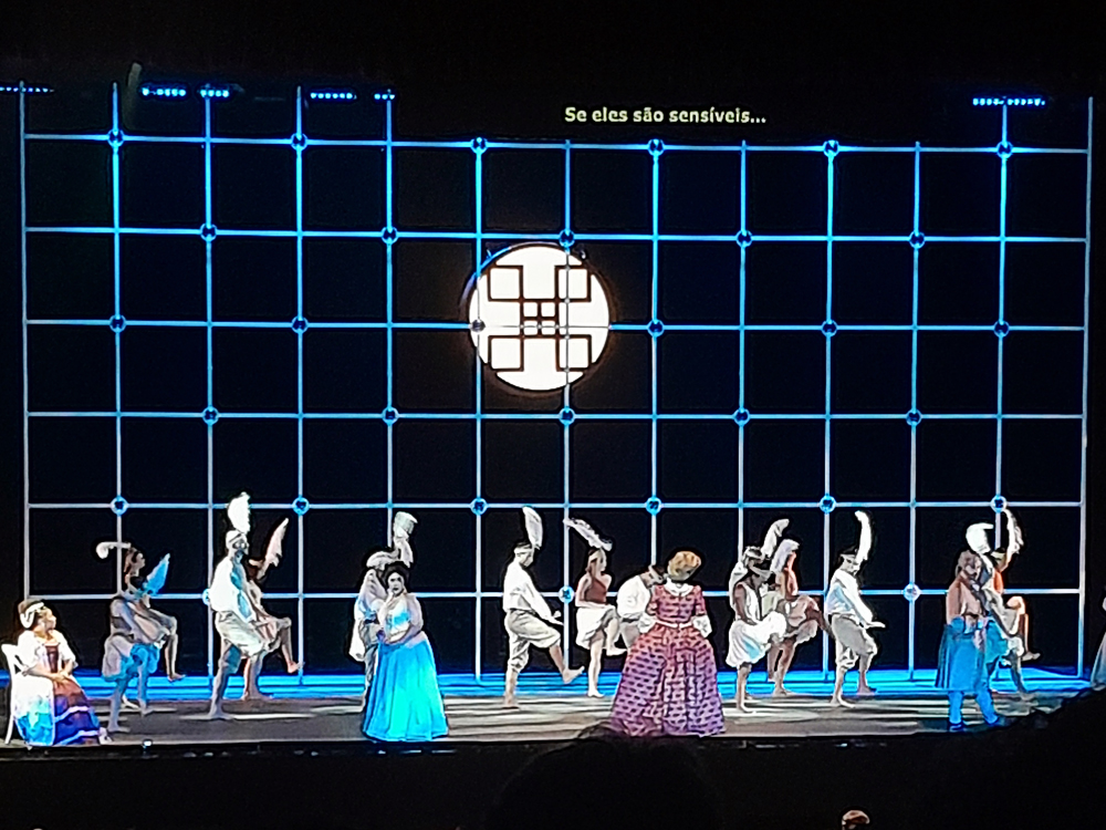 Conjunto de artistas ocupa o palco em formação ampla. Alguns usam cocares com penas, outros vestidos coloridos, compondo quadro vibrante. O fundo azul quadriculado tem círculo luminoso central, evocando lua ou janela. A legenda projetada acompanha o canto, enquanto a coreografia dá ritmo à narrativa.