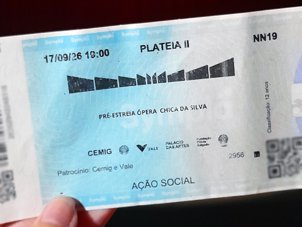 Mão segura ingresso azul da pré?estreia da ópera Chica da Silva no Palácio das Artes, com informações de data, horário, setor, classificação e logos de patrocinadores como Cemig e Vale. 