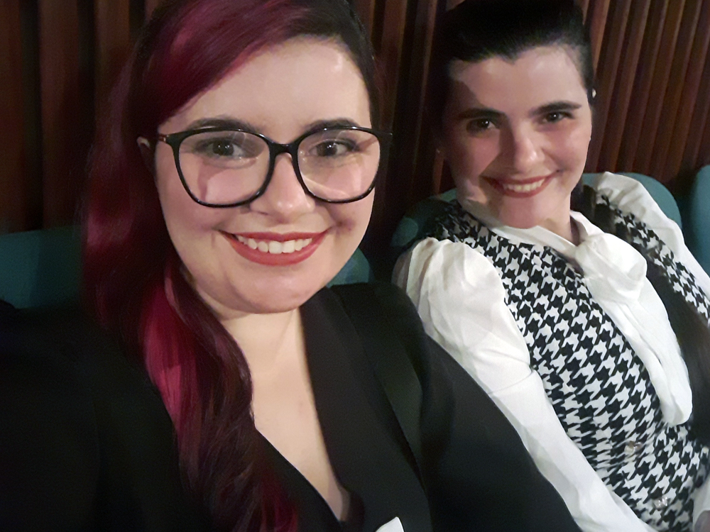 Duas mulheres sentadas em poltronas internas. À esquerda, pessoa de cabelos rosa escuros, óculos e roupa preta. À direita, pessoa de cabelos escuros presos, usando blusa branca com colete xadrez preto e branco. Ambas olham para a câmera em ambiente de teatro com madeira ao fundo.