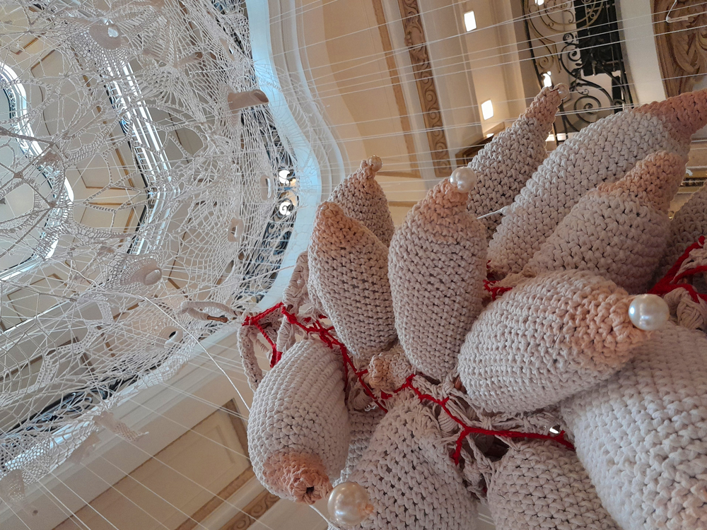 Instalação da exposição Tecitura do feminino, de Marlene Barros, vista de baixo, com conjunto de formas orgânicas volumosas semelhantes a seios feitas em crochê branco e cru, algumas alongadas e pontiagudas e outras arredondadas. Pérolas brancas aparecem aplicadas em várias extremidades, enquanto fios e pequenos trechos vermelhos atravessam a composição. À esquerda, uma grande estrutura circular vazada, semelhante a uma teia de renda, ocupa parte do espaço e deixa visível a arquitetura do edifício ao fundo.