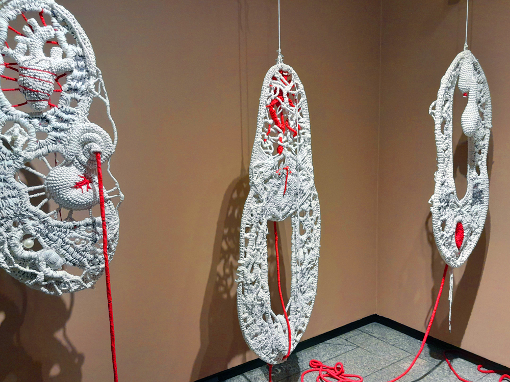 Três estruturas alongadas de crochê branco suspensas verticalmente diante de uma parede bege. As peças são vazadas e irregulares, formadas por diferentes pontos, tramas e volumes que lembram partes de uma anatomia abstrata. Fios de vermelho intenso atravessam o crochê, descem das estruturas e se acumulam parcialmente sobre o piso, criando forte contraste com o branco das fibras.