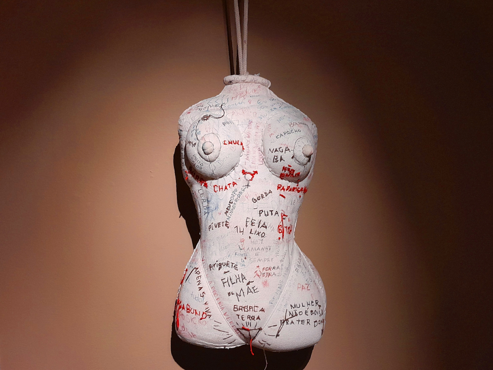 Escultura suspensa de um torso revestido por tecido branco, coberto por palavras e expressões manuscritas e bordadas em preto, cinza e vermelho. Termos como chata, gorda, puta, feia, filha, mãe, barata, vagabunda e mulher se espalham pelo corpo, acompanhados por linhas e marcas de costura. A obra está iluminada frontalmente contra uma parede marrom, projetando uma sombra escura atrás da figura.