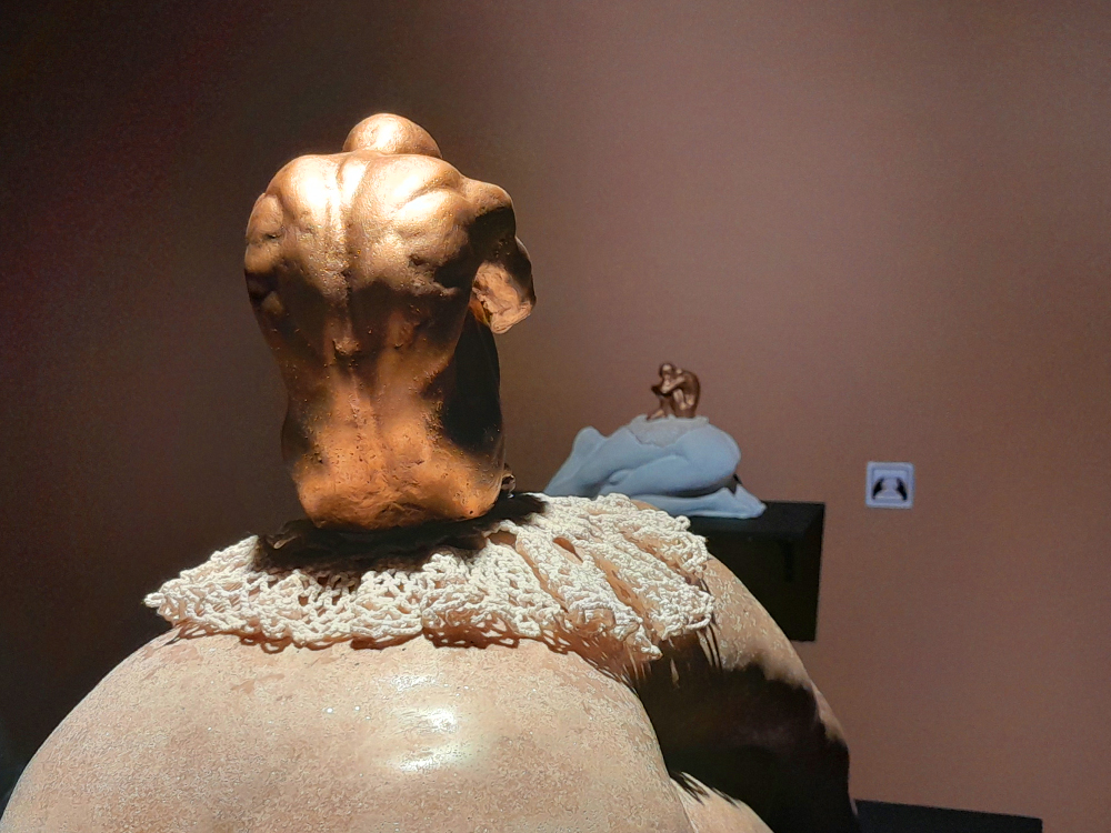 Escultura de um torso de costas com cabeça abaixada, em tom dourado e superfície irregular, apoiado sobre uma grande forma corporal arredondada coberta parcialmente por uma peça de crochê branco semelhante a uma gola de renda. A iluminação lateral acentua os volumes e as texturas da escultura. Ao fundo, sobre uma prateleira preta, aparece uma versão tátil da obra em escala reduzida, com as formas principais reproduzidas em materiais de cores e texturas contrastantes; ao lado, um símbolo indica o recurso de acessibilidade tátil.