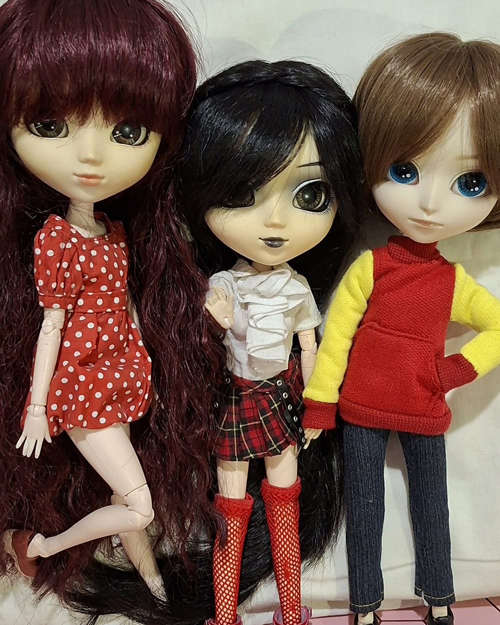 Três bonecas articuladas posam lado a lado sobre fundo claro. À esquerda, uma Pullip de cabelos longos vinho usa vestido vermelho com bolinhas brancas. No centro, uma Pullip de cabelos pretos longos veste blusa branca com babados, saia xadrez vermelha e preta e meias arrastão vermelhas até os joelhos. À direita, um Isul de cabelos castanhos e olhos azuis usa blusa vermelha com mangas amarelas e calça jeans escura.