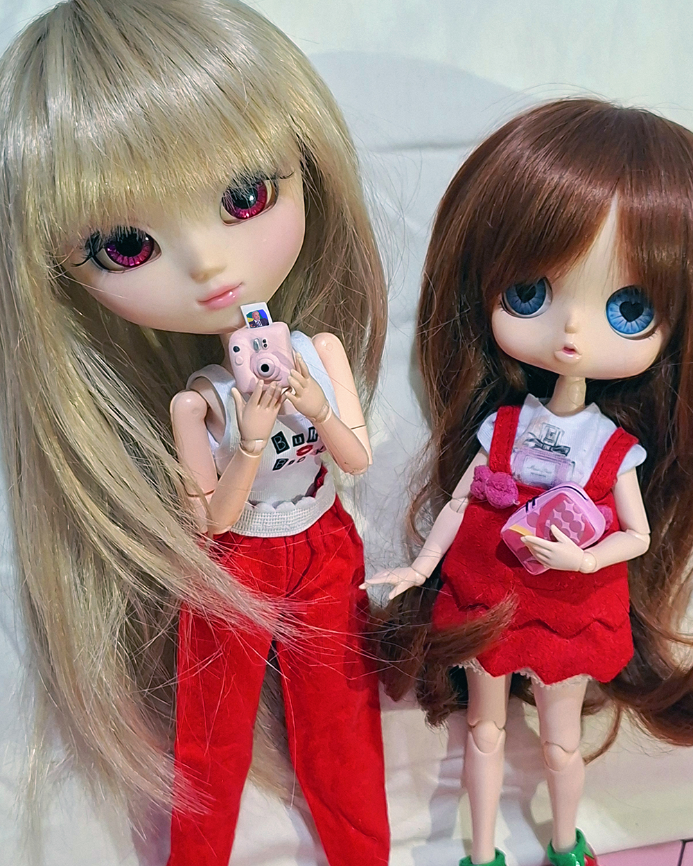 Duas bonecas articuladas posam lado a lado sobre fundo claro. À esquerda, uma Pullip de cabelos loiros muito longos e olhos rosa segura uma câmera instantânea rosa com uma fotografia do Lula saindo da parte superior. Ela veste camiseta branca e calça vermelha. À direita, uma boneca Byul, menor, de cabelos castanhos ondulados e olhos azuis usa vestido vermelho sobre camiseta branca e segura uma pequena bolsa rosa estampada.