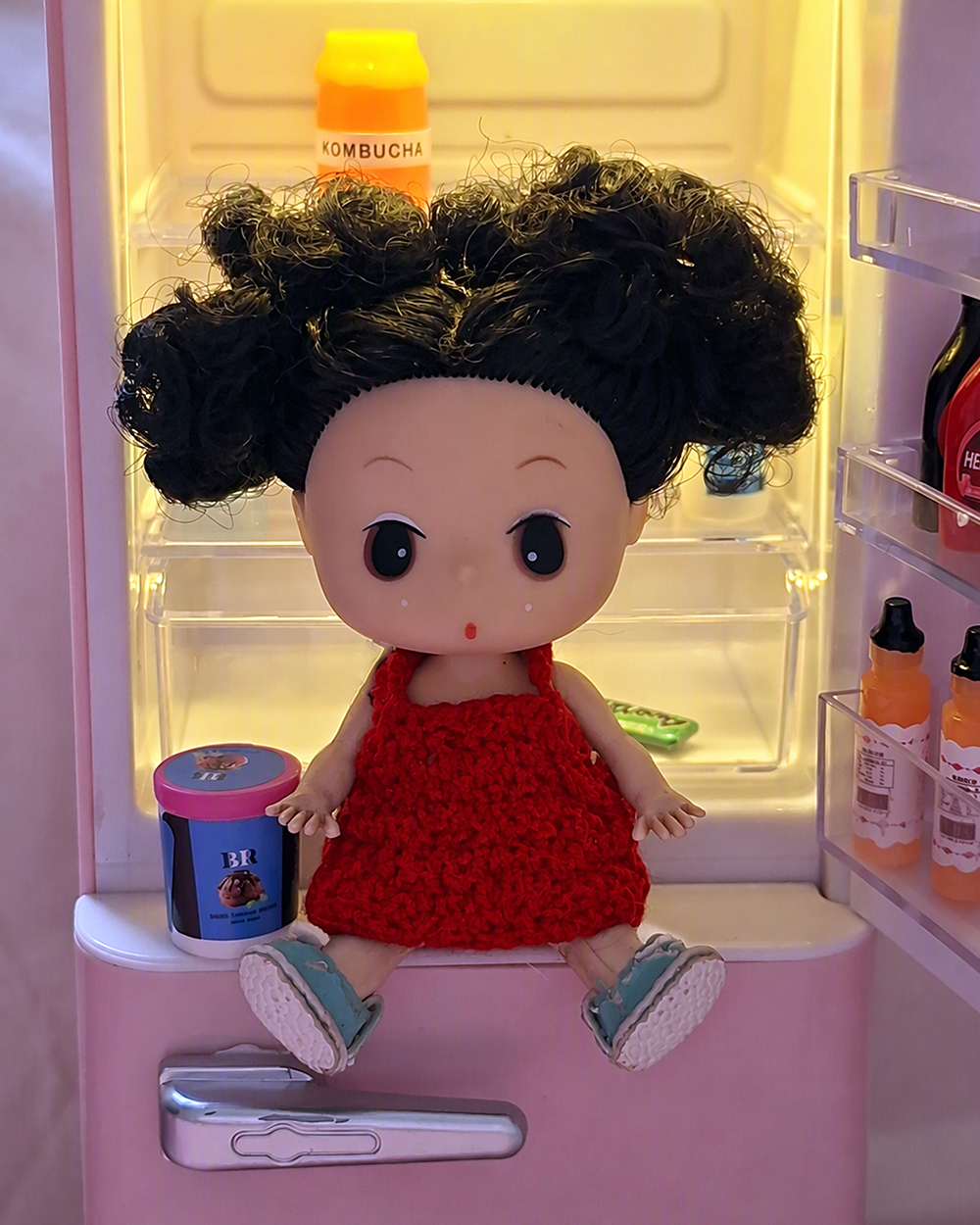 Boneca em estilo dDung sentada na porta aberta de uma mini geladeira rosa iluminada por luz amarela. A boneca tem cabelos pretos encaracolados presos em dois coques laterais e veste vestido vermelho de tricô com tênis azul-claro. Ao lado, sobre a porta da geladeira, há um pequeno pote de sorvete azul e rosa. No interior aparecem uma garrafa identificada como kombucha, pequenas embalagens e miniaturas de alimentos organizadas nas prateleiras transparentes.