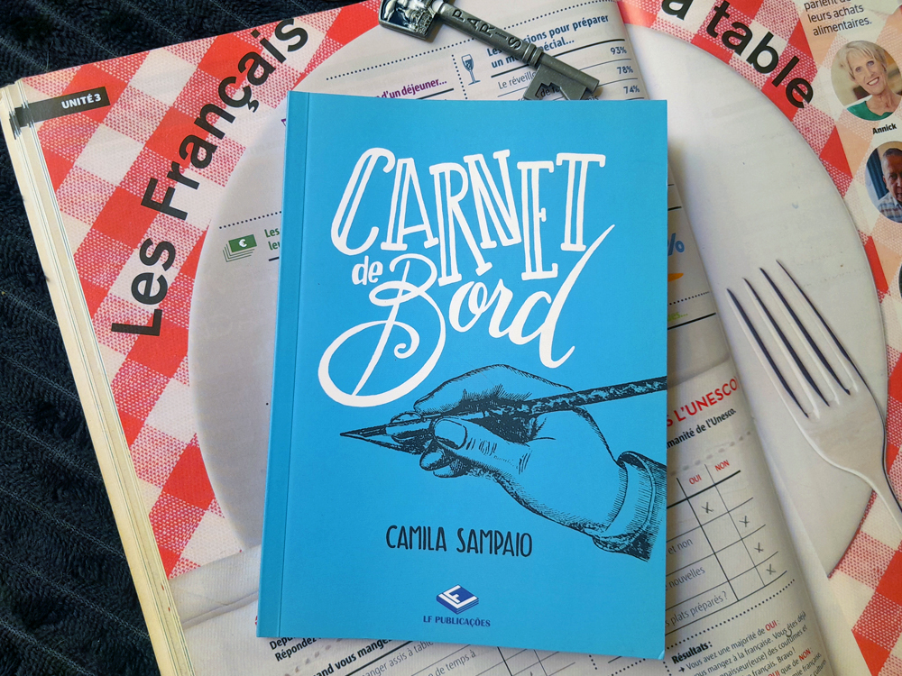 Livro Carnet de bord, de Camila Sampaio, apoiado sobre páginas de uma revista didática em francês com estampa de toalha xadrez vermelha e branca e fotografia de prato e garfo. A capa azul traz o título em letras brancas, uma mão segurando uma caneta e o nome da autora. Uma pequena chave de Paris aparece acima do livro.