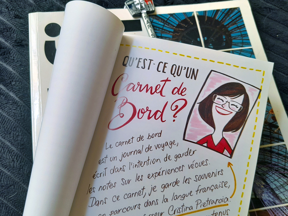 Livro Carnet de bord, de Camila Sampaio, aberto em uma página com o título em francês “Qu’est-ce qu’un Carnet de Bord?”. A página traz texto escrito à mão e uma ilustração de pessoa de cabelos castanhos e óculos. Ao fundo aparecem outros livros e uma pequena chave metálica de Paris.