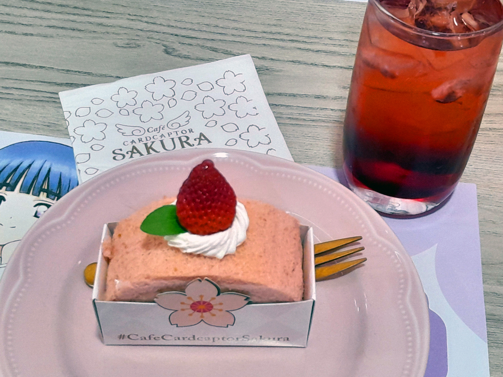 Sobremesa rosa de cereja servida em prato também rosa, decorada com chantilly, morango inteiro e folha verde. Ao lado, copo com bebida vermelha e gelo. Um cartão ao fundo traz flores de cerejeira e o nome Café Cardcaptor Sakura, enquanto parte de uma ilustração de personagem aparece sob o prato.