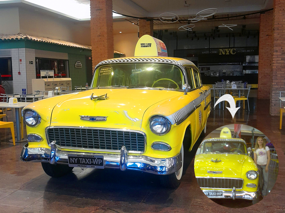 Automóvel Chevrolet antigo transformado em táxi amarelo de inspiração nova-iorquina, estacionado dentro de um espaço com mesas e restaurantes. O carro tem detalhes cromados, faixa quadriculada preta e branca, identificação “Yellow” no teto e placa decorativa “NY TAXI-WP”. No canto inferior direito, um recorte circular mostra uma fotografia antiga do mesmo carro ao lado de uma adolescente loira, ligado à imagem principal por uma seta cinza.