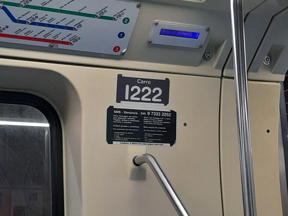 Interior de um vagão do metrô, com placa identificando o carro número 1222 acima de um aviso com telefone para denúncias por SMS. Na parte superior aparecem um mapa das linhas do metrô e um pequeno painel eletrônico azul; barras metálicas e parte da porta completam a cena.